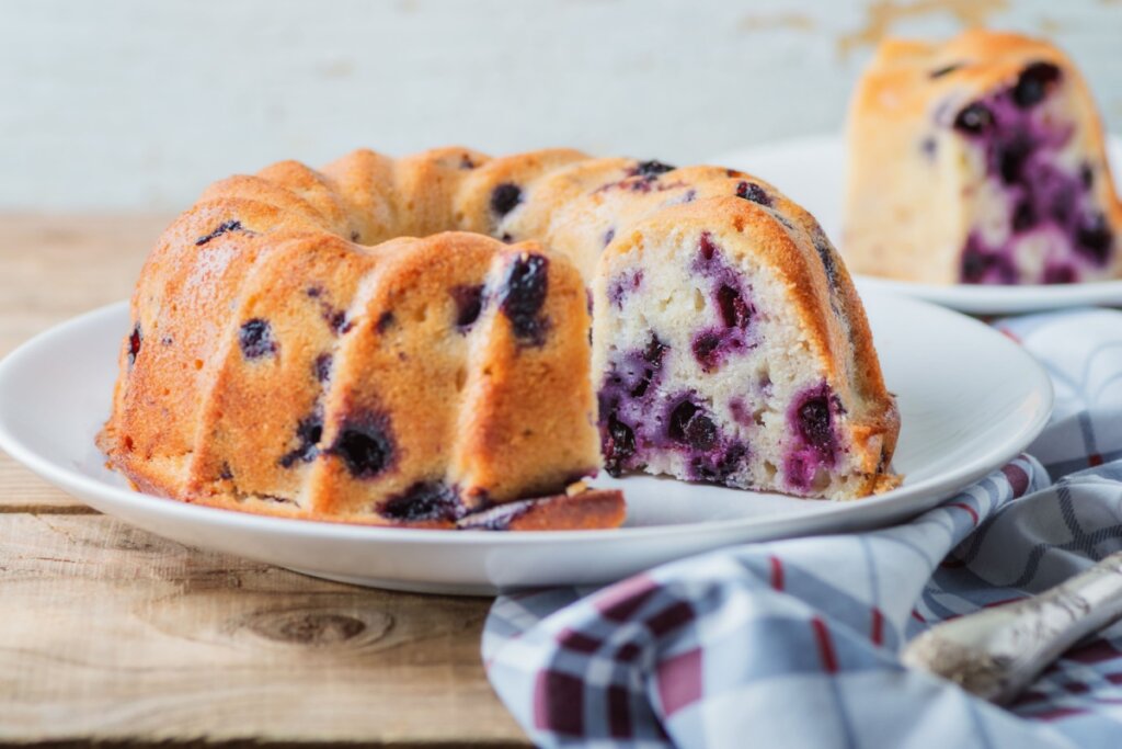 Bolo de iogurte com mirtilo em cima de um prato branco e com uma das fatias cortadas 