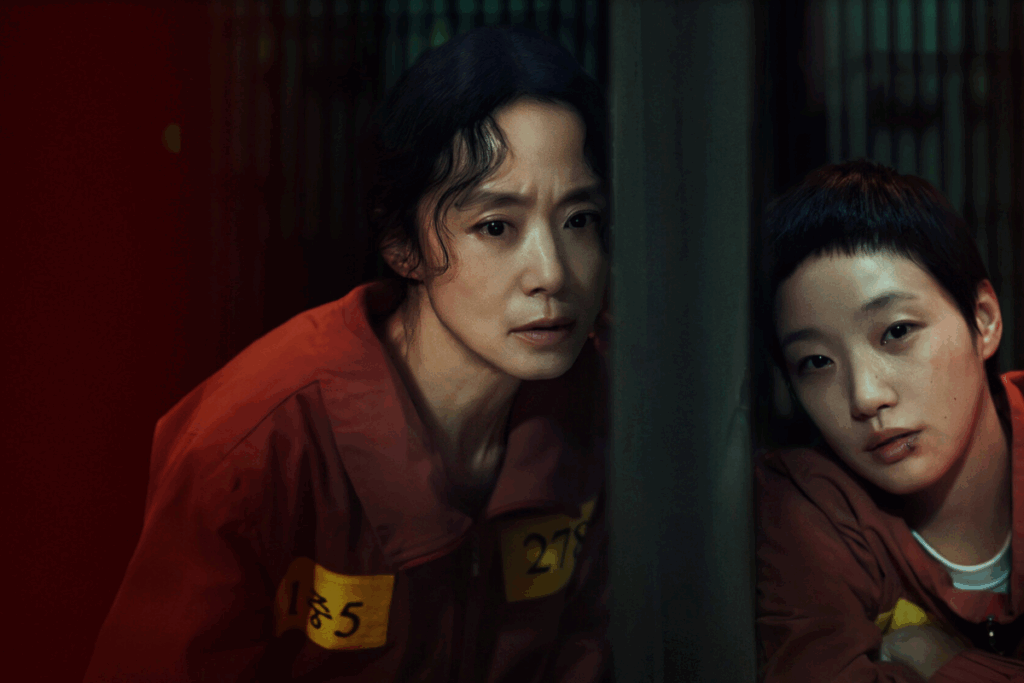 Duas mulheres usando uniforme de detentas encostadas em parede de cela