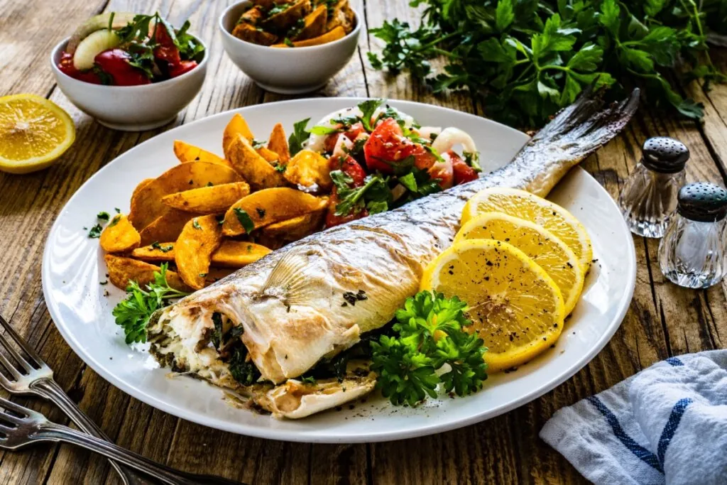 Peixe inteiro assado com rodelas de limão, batatas douradas e salada fresca colorida servidos em prato branco sobre mesa de madeira