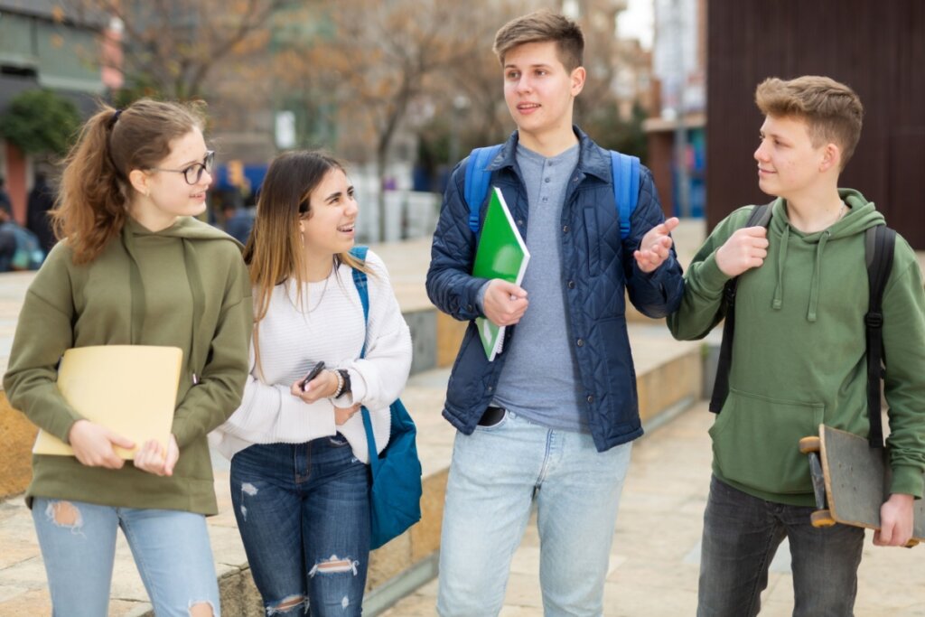 Adolescentes andando na rua, segurando livros e mochilas, e conversando 