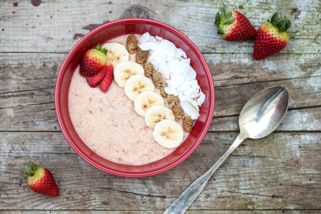 Recipiente vermelho com mingau de aveia com morango, decorado com rodelas de banana, lascas de coco e nozes