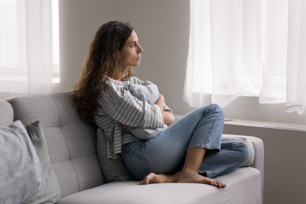 mulher de camiseta listrada, calça jeans, sentada em sofá cinza, abraçada em almofada e olhando para o lado