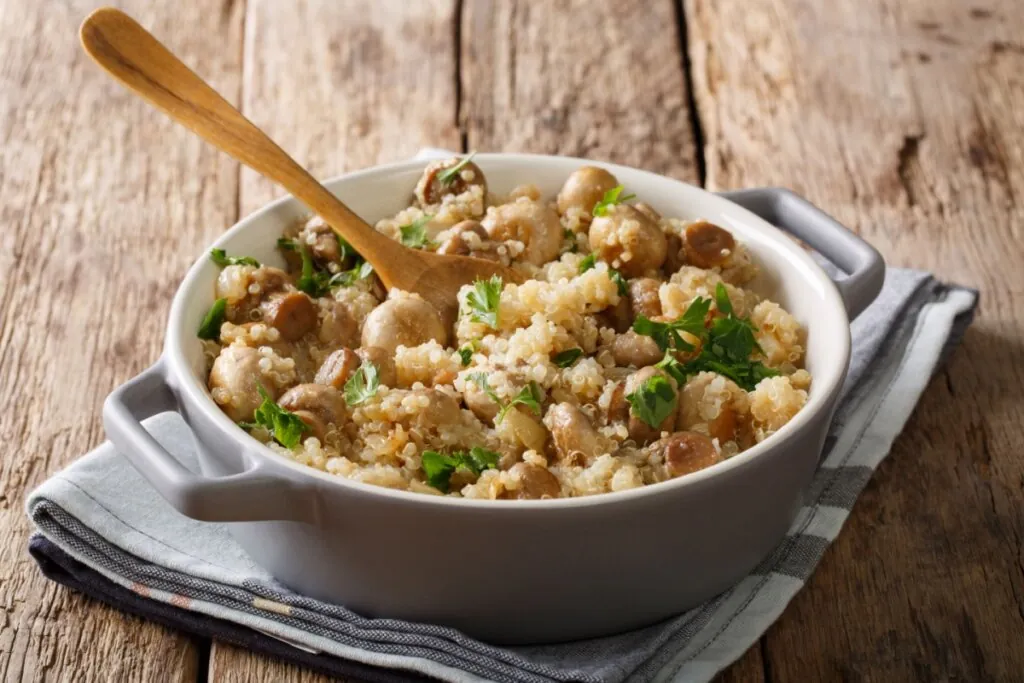 Quinoa com cogumelos salteados servido em panela de cerâmica branca em cima de pano de prato listrado em cima de mesa de madeira
