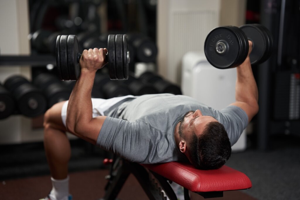 Homem realizando exercício de supino reto com halteres em banco de academia
