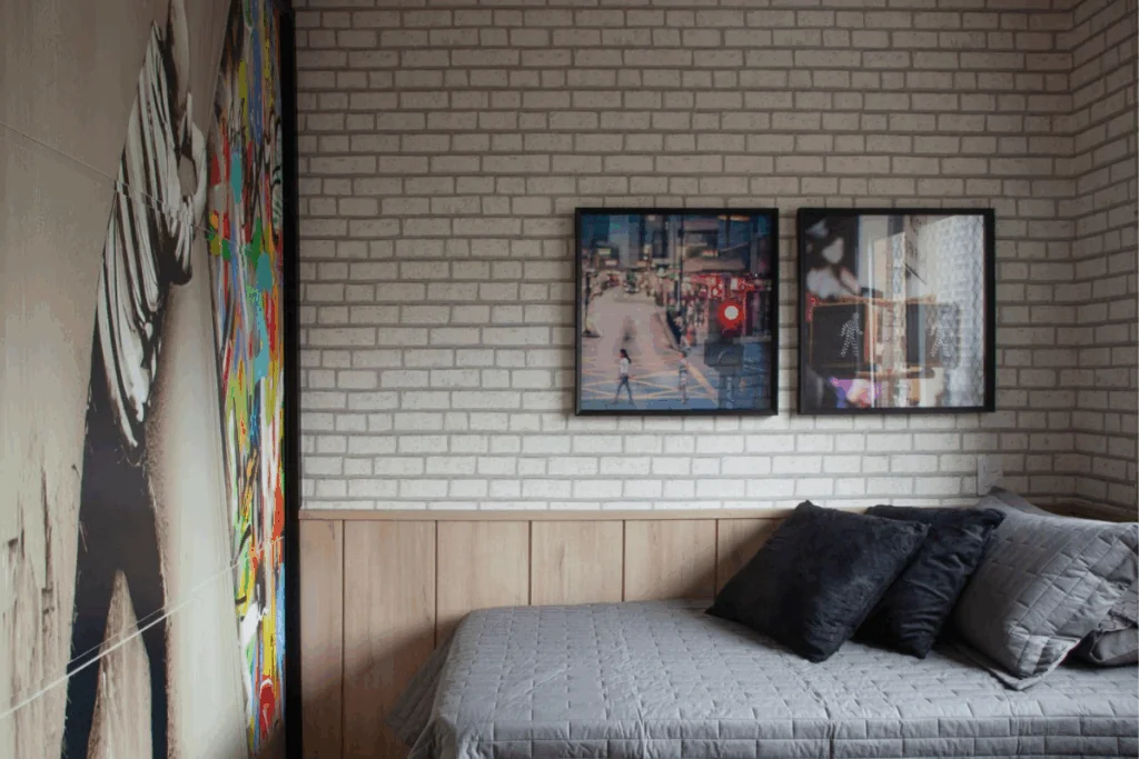 Quarto com papel de parede com estampa de tijolos, com dois quadros e na outra parede um papel de parede com um homem simulando que está rasgando a parede em colorido e cama de solteiro com colcha cinza e almofadas cinza e preta