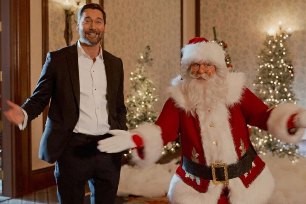 Homem vestindo roupa social preta e ao lado de um papai noel, ambos sorrindo e em pé 