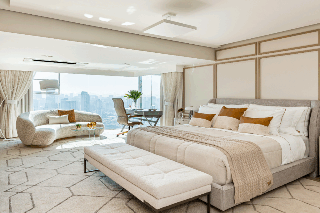 Parede bege com boiserie dourada em quarto com cama de casal com roupa de cama bege e travesseiros com capa bege e marrom com parede de fundo de vidro e sofá claro e mesa de escritório