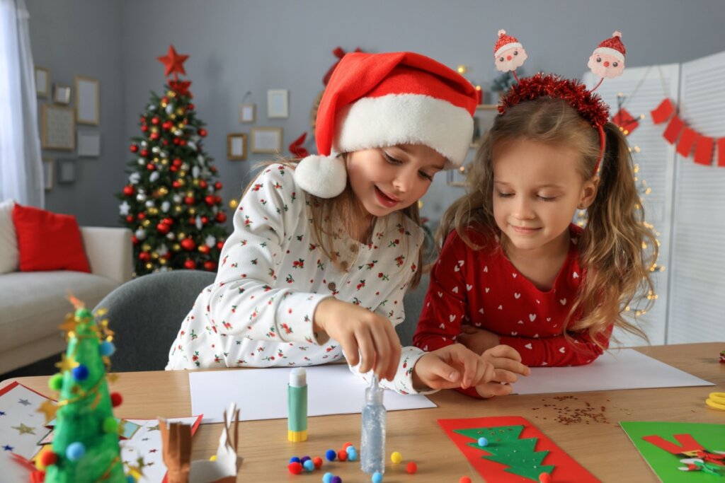 Duas crianças de pijama natalino fazendo cartões juntas em uma mesa
