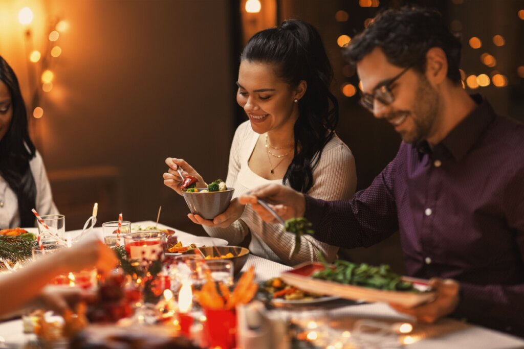 Homem e mulher sentados em uma mesa com ceia de Natal, sorrindo e colocando alimentos no prato 