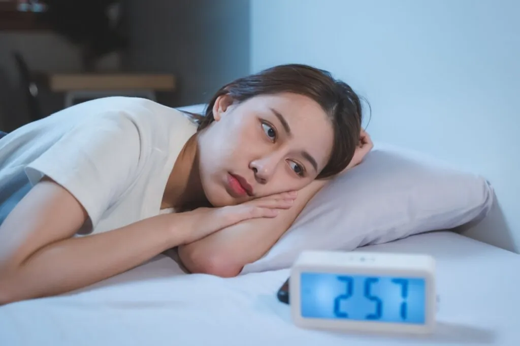 Jovem deitada na cama, usando camiseta branca sem conseguir dormir e a frente no móvel ao lado da cama um relógio marcando 2h57 da manhã