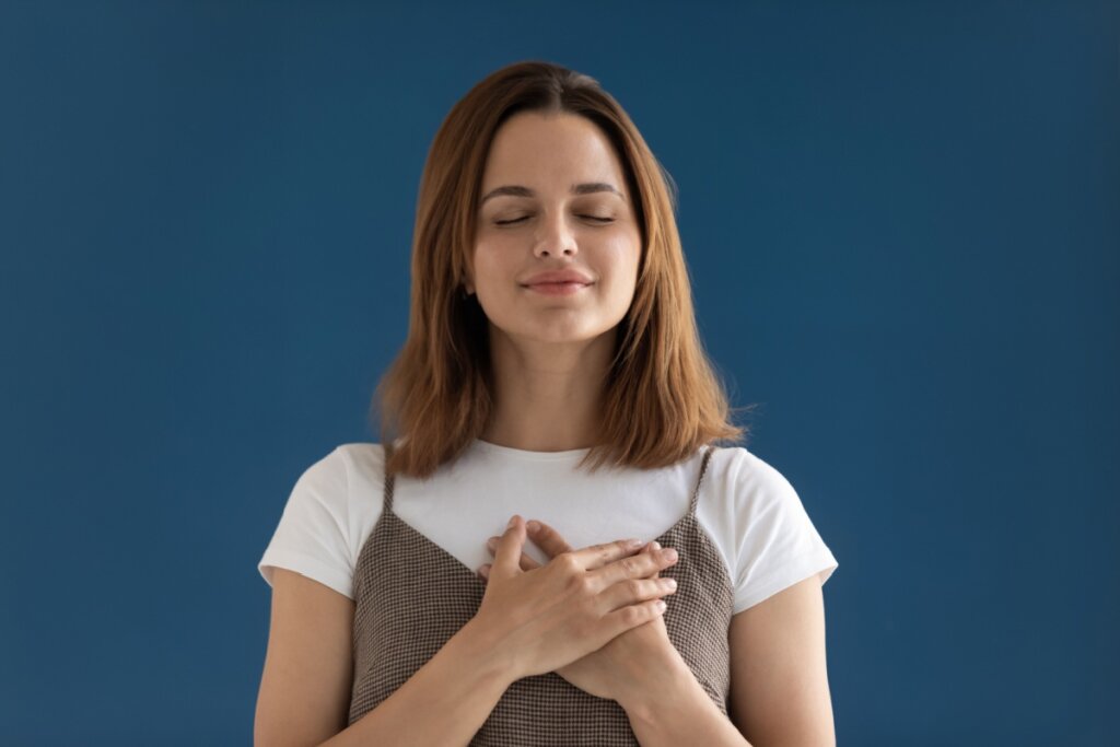 mulher de blusa branca e marrom, com os olhos fechados, sorrindo e com as duas mãos no peito