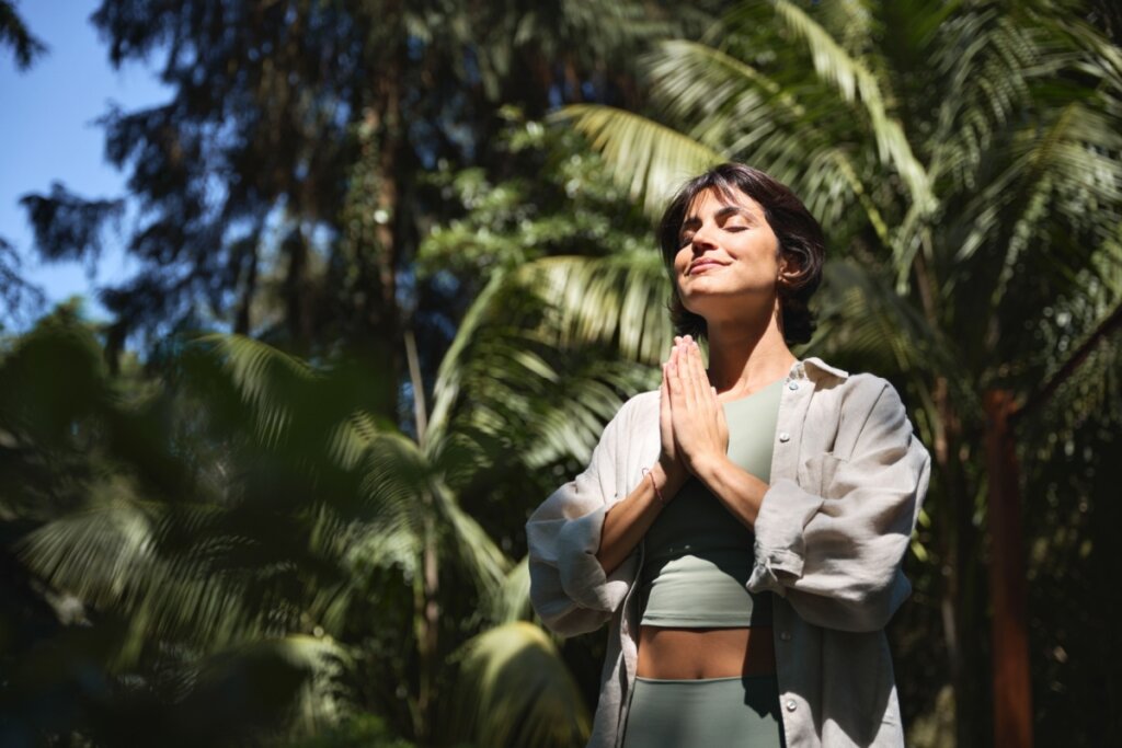 mulher de roupa verde e cinza, com cabelo curto, olhos fechados e mãos unidas, rezando. árvores ao fundo