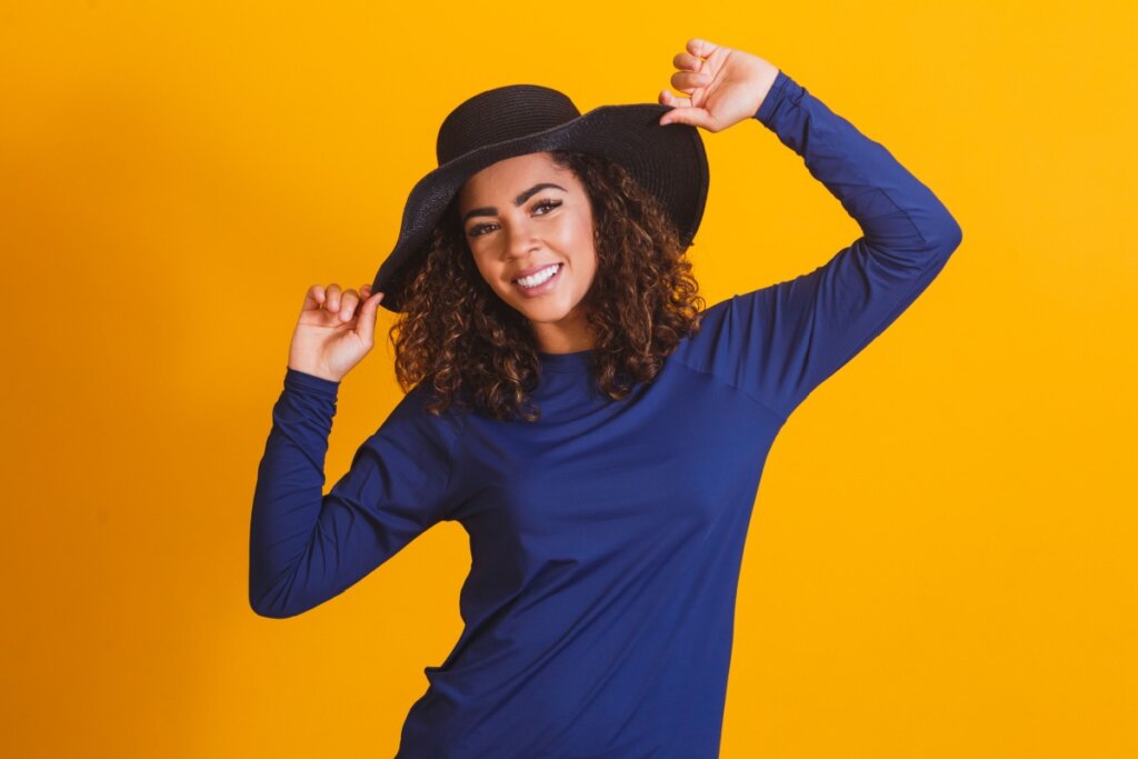 Mulher com cabelo cacheado usando chapéu preto, e camisa de manga longa com proteção UV azul marinho em fundo laranja