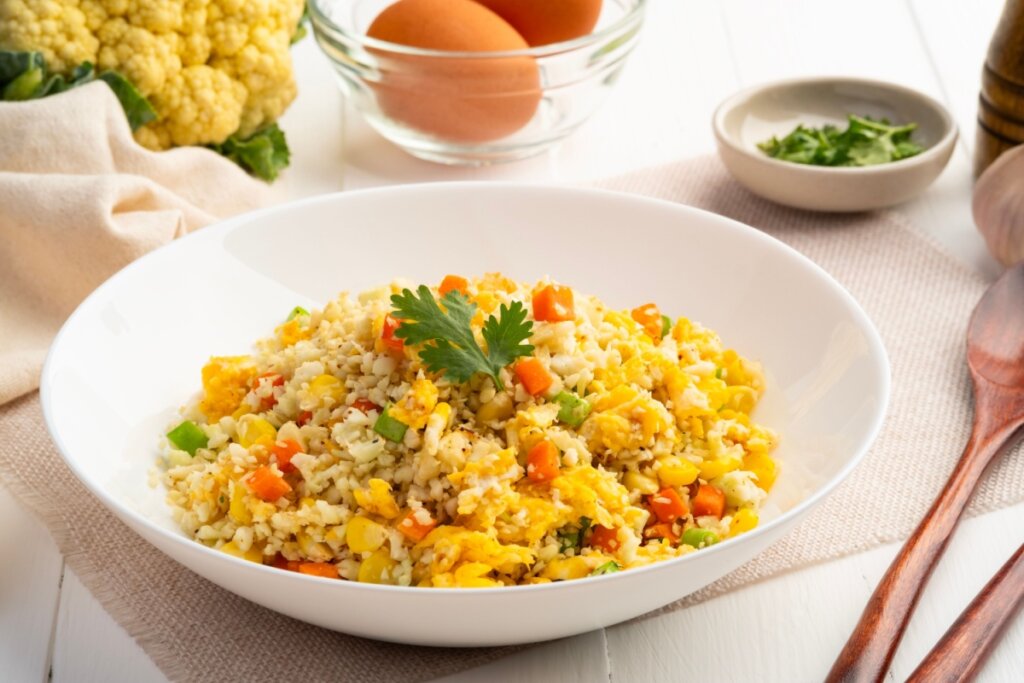 Prato com arroz de couve-flor misturado com ovos mexidos, cenoura em cubos e milho, servido em prato branco fundo