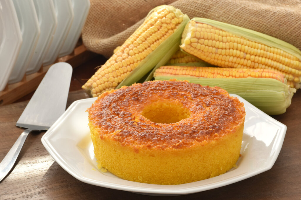Bolo de milho em cima de um prato branco ao lado de três unidades de milho-verde