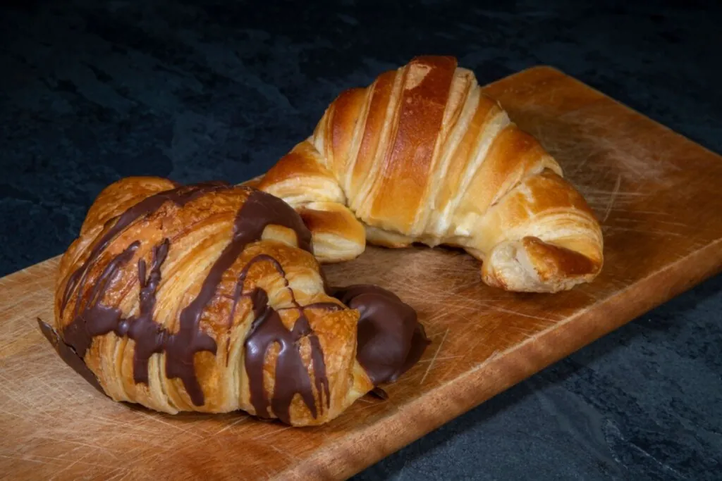 Dois croissants grandes dispostos em uma tábua de madeira. O croissant em primeiro plano está coberto com listras de chocolate derretido e possui um recheio de chocolate cremoso saindo pela lateral. O segundo, ao fundo, é simples e mostra bem as camadas douradas e curvas da massa folhada tradicional.