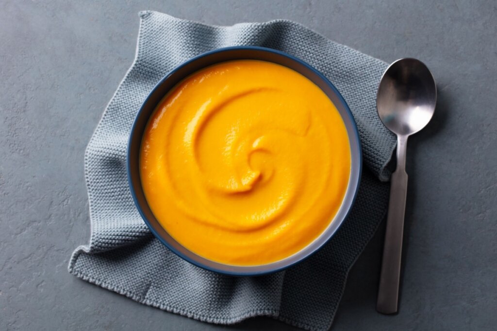 Purê de lentilha com batata-doce em uma tigela. A tigela está posicionada sobre um pano de prato de tecido cinza-azulado com textura de colmeia. Ao lado direito, há uma colher de metal fosco disposta verticalmente.