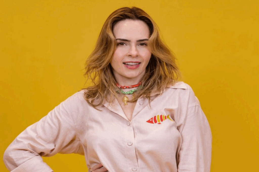 Mulher com cabelo curto com mechas loiras usando macacão rosa-claro, com mão na cintura em fundo amarelo