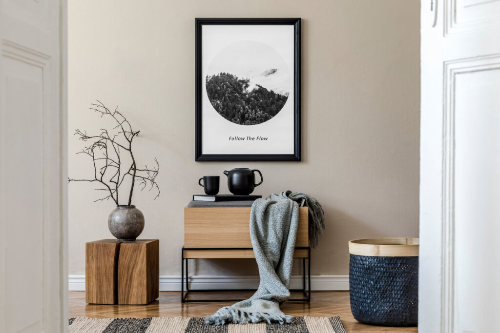 Parede bege com quadro preto e branco, abaixo, mesas de madeira com bule preto, vaso e manta cinza 