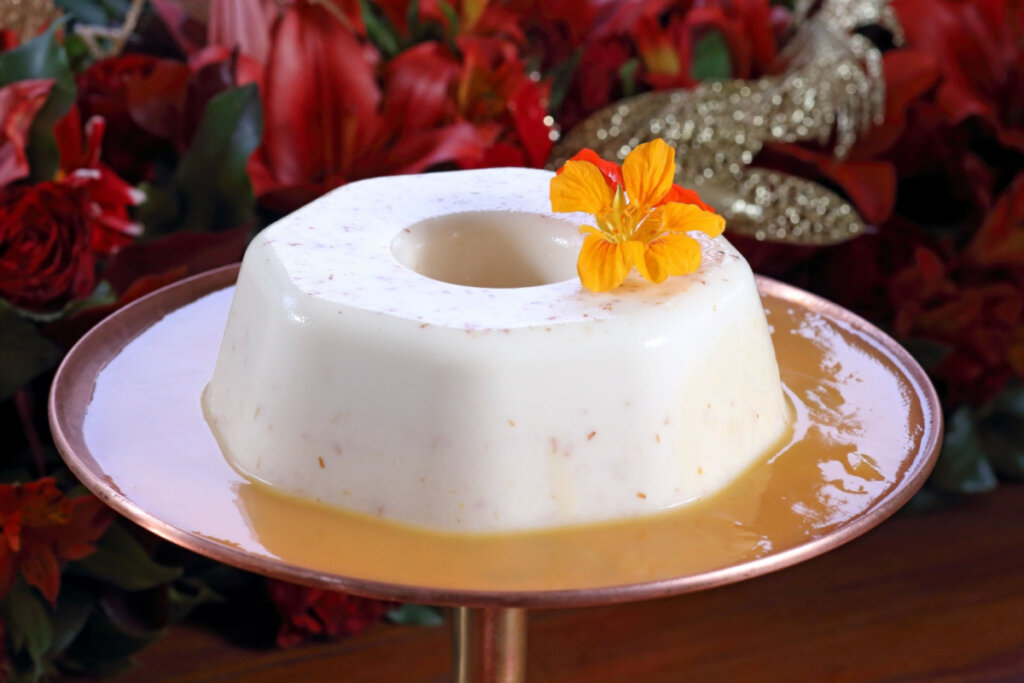 Pudim de coco desenformado sobre prato com calda clara, decorado com uma flor amarela