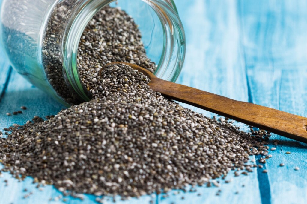 Recipiente de vidro deitado em uma mesa de madeira azul com sementes de chia saindo dele e uma colher de madeira dentro