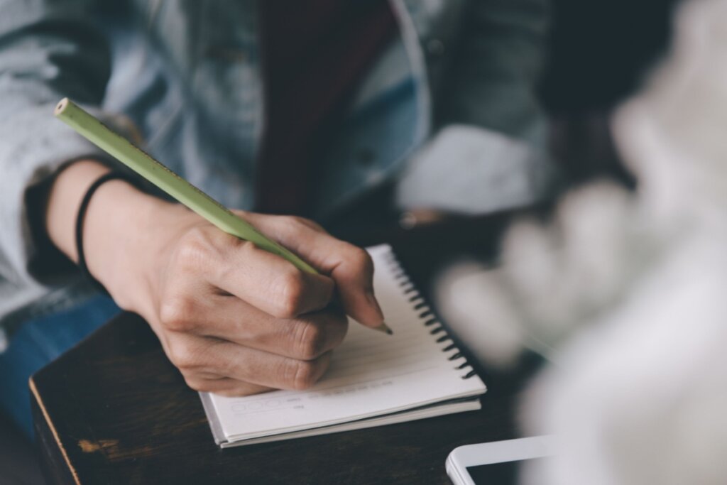 Mão segurando um lápis verde e escrevendo em um caderno com folhas brancas 