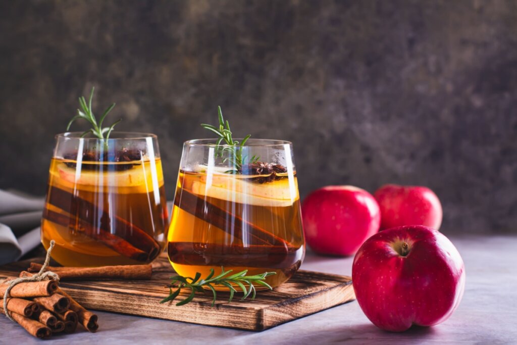 Dois copos de vidro com chá claro, maçã, canela e alecrim sobre tábua de madeira, acompanhados por maçãs inteiras e paus de canela ao redor