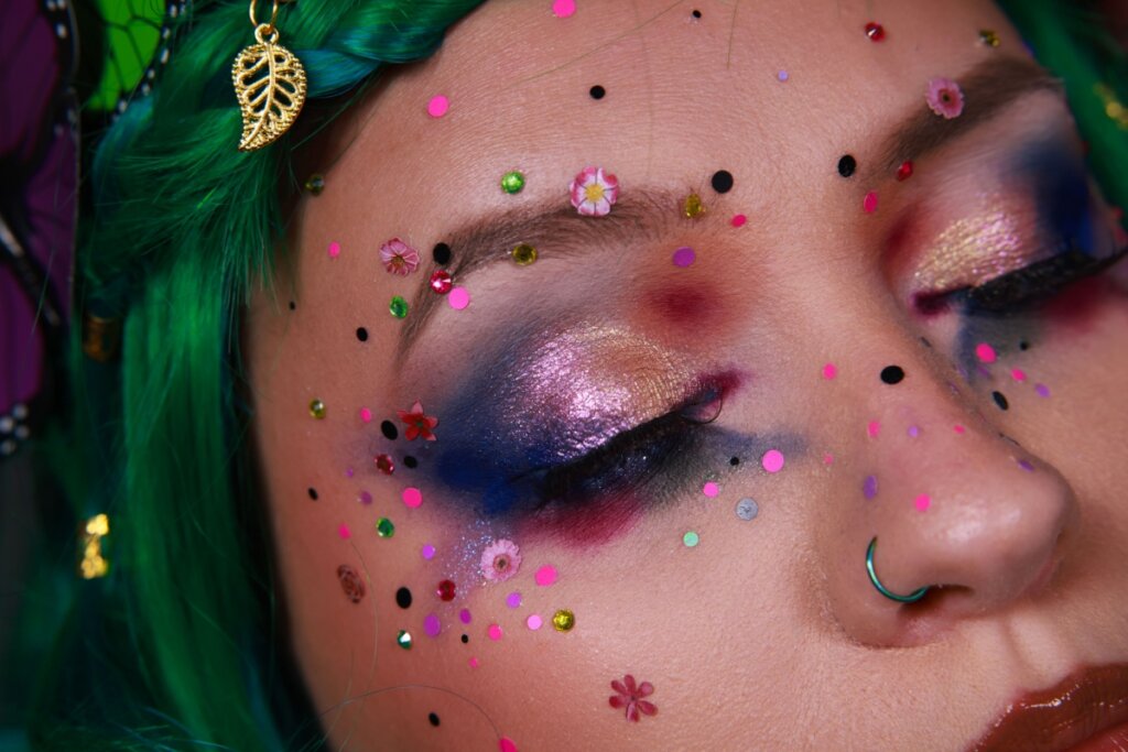 Fotografia em close-up do rosto de uma pessoa com uma maquiagem artística e maximalista. Os olhos estão fechados, exibindo uma sombra cintilante em tons de rosa, dourado e azul profundo. O rosto está decorado com confetes coloridos, pequenos cristais brilhantes e miniaturas de flores coladas na pele. O cabelo é de um verde vibrante com uma trança, adornado por um pingente de folha dourada. A pessoa também possui um piercing de argola azul no nariz e lábios com gloss marrom, criando um visual lúdico e fantasioso.