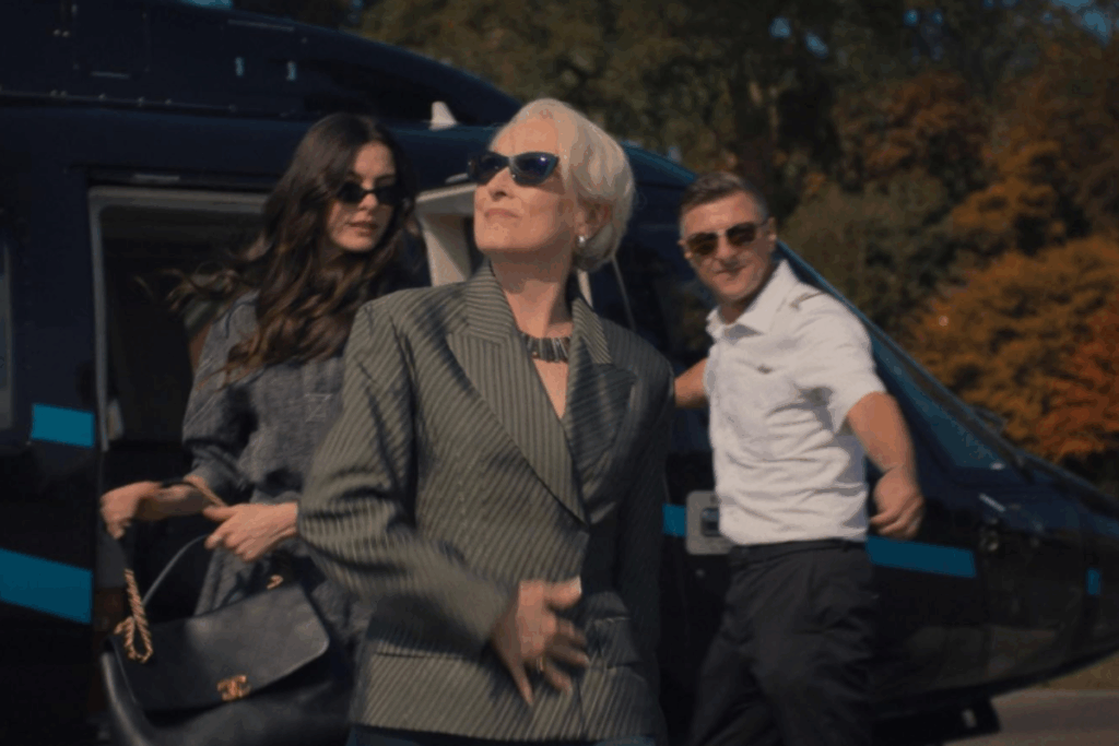 Uma cena do filme "O Diabo Veste Prada 2". Em primeiro plano, Miranda Priestly (Meryl Streep) caminha à esquerda, usando um blazer cinza estruturado, óculos de armação escura e um colar de corrente prateada, mantendo sua expressão icônica e autoritária. À direita, Nigel (Stanley Tucci) caminha ao seu lado, vestindo um terno xadrez bege elegante e óculos redondos. Entre e ligeiramente atrás deles, Andy Sachs (Anne Hathaway) aparece sorrindo, com longos cabelos escuros e um visual sofisticado composto por um vestido azul-marinho e um colar de pérolas. O trio caminha com confiança por um escritório de luxo.