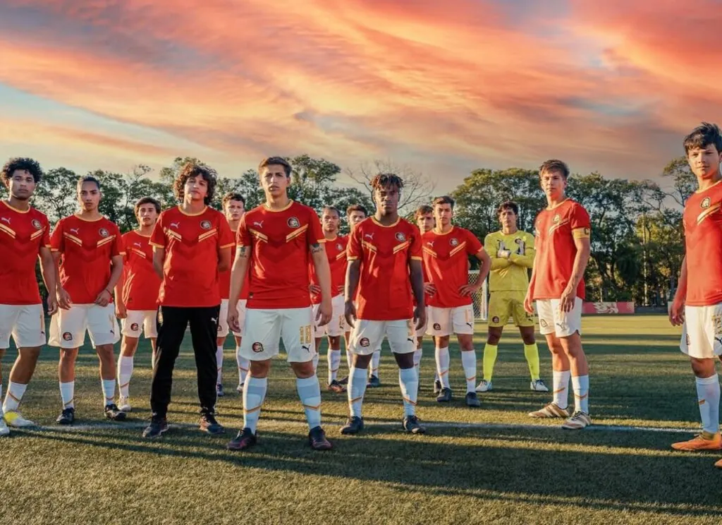 Foto de grupo de um time de futebol juvenil posando em um campo de grama sintética durante o pôr do sol. Os jogadores vestem uniformes vermelhos com detalhes em amarelo e calções brancos. Eles estão espalhados pelo campo com expressões confiantes. O céu ao fundo apresenta nuvens em tons vibrantes de laranja e rosa, criando um clima épico e inspirador.