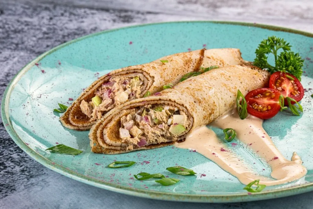Duas panquecas cortadas ao meio, revelando um recheio cremoso de atum com cebola roxa e cebolinha. Estão servidas em um prato azul decorado com molho rosado, tomates cereja.