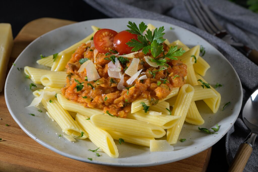 Prato de penne com molho bolonhesa de lentilha, finalizado com tomate-cereja e salsinha e servido sobre tábua de madeira