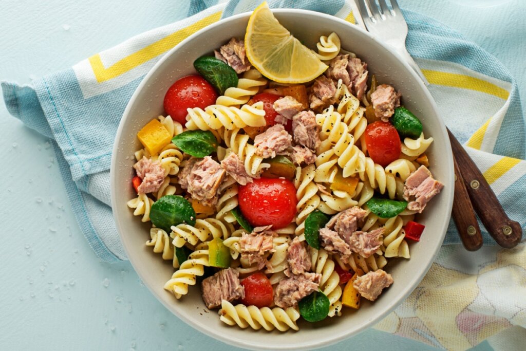 Recipiente branco com macarrão com atum e vegetais em cima de uma mesa forrada com pano azul-claro