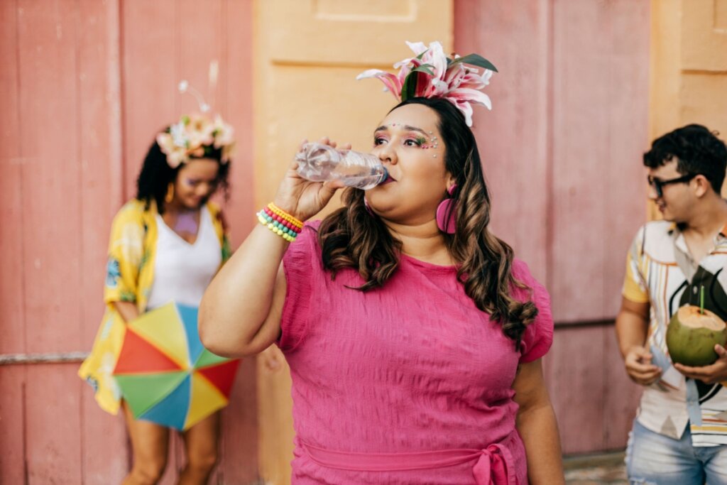 Mulher com cabelo longo, castanho e solto, usando tiara com penas coloridas, brinco grande rosa, pulseiras coloridas e camiseta rosa tomando água de garrafa plástica em bloco de Carnaval