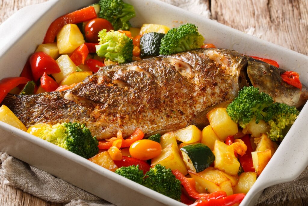 Peixe assado com legumes servido em travessa de cerâmica branca em cima de mesa de madeira