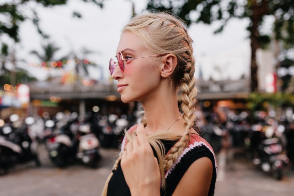 Mulher com trança embutida no cabelo, virada de lado, e usando um óculos de sol rosa 