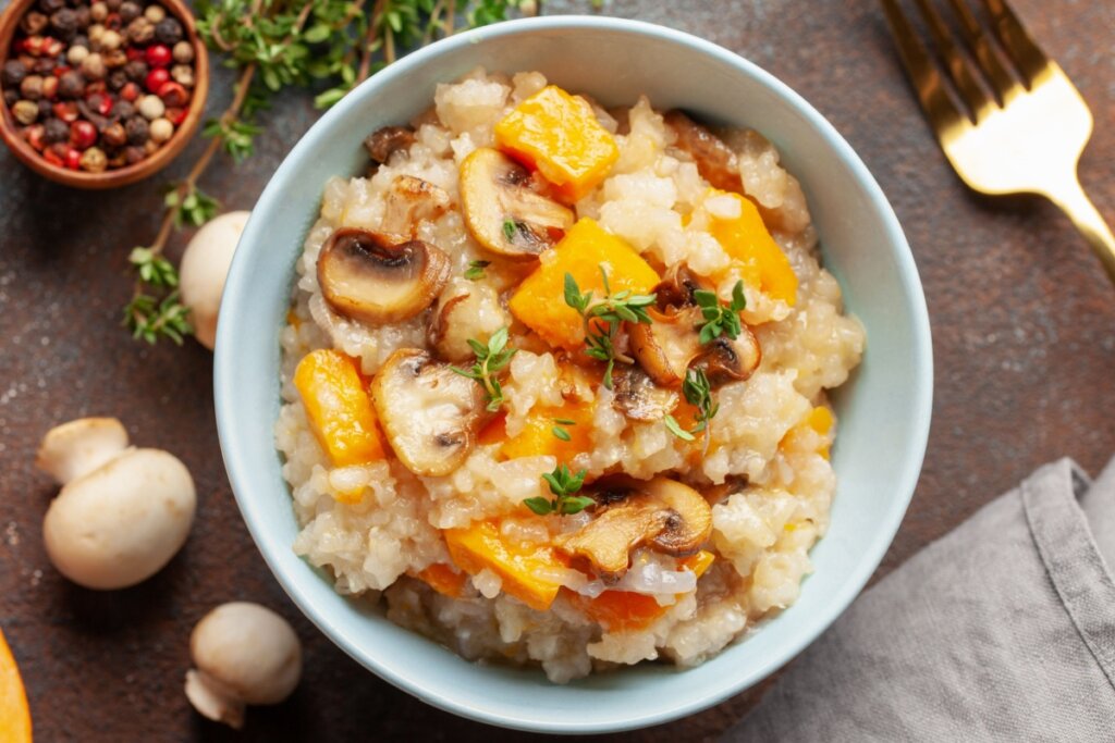 Risoto de abóbora com cogumelo servido em prato fundo em cima de mesa de madeira, com garfo de ferro ao lado e cogumelos