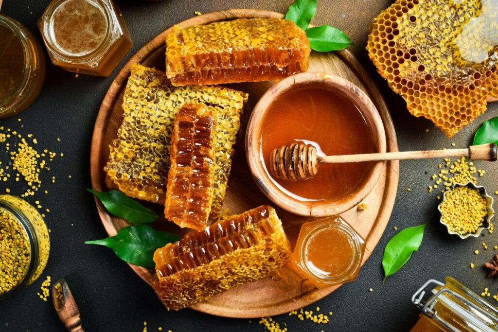 Vista superior de uma mesa composta por diversos elementos apícolas. No centro, uma tábua redonda de madeira contém vários pedaços de favo de mel e uma tigela pequena com mel líquido e um pegador. Ao redor, há potes de vidro com mel, grãos de pólen espalhados, folhas verdes e favos inteiros sobre um fundo escuro texturizado.