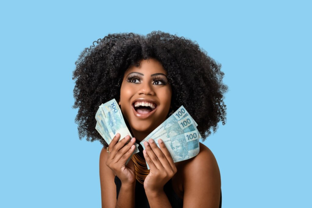Uma mulher negra com cabelo crespo volumoso sorri alegremente e olha para cima, segurando vários leques de notas de 100 reais próximos ao rosto. Ela usa uma blusa preta e um colar dourado, destacando-se contra um fundo azul sólido. A imagem transmite uma sensação de sucesso financeiro, felicidade e prosperidade.