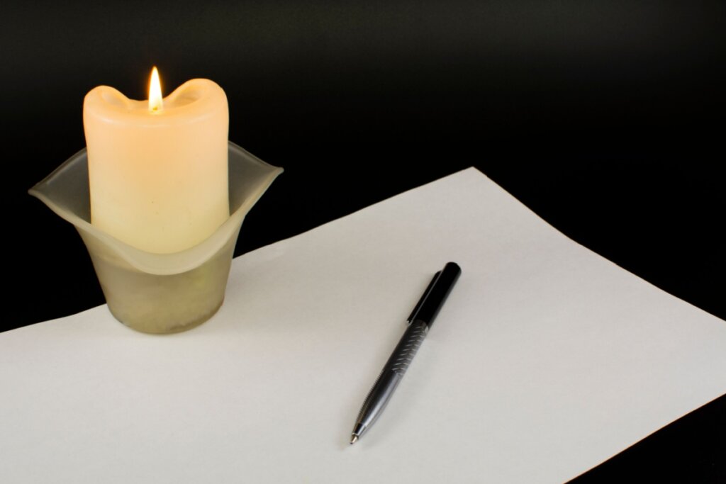 Uma folha de papel em branco com uma caneta preta e prateada sobre ela. À esquerda, uma vela grossa de cor branca está acesa dentro de um suporte de vidro fosco, com a chama brilhando contra um fundo preto sólido. A cena sugere um momento de escrita introspectiva, ritual ou reflexão.