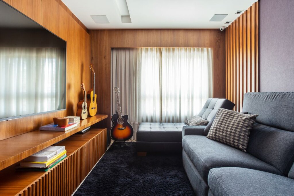 Sala de estar com televisão, sofá cinza e, ao fundo, janela com cortina blackout branca