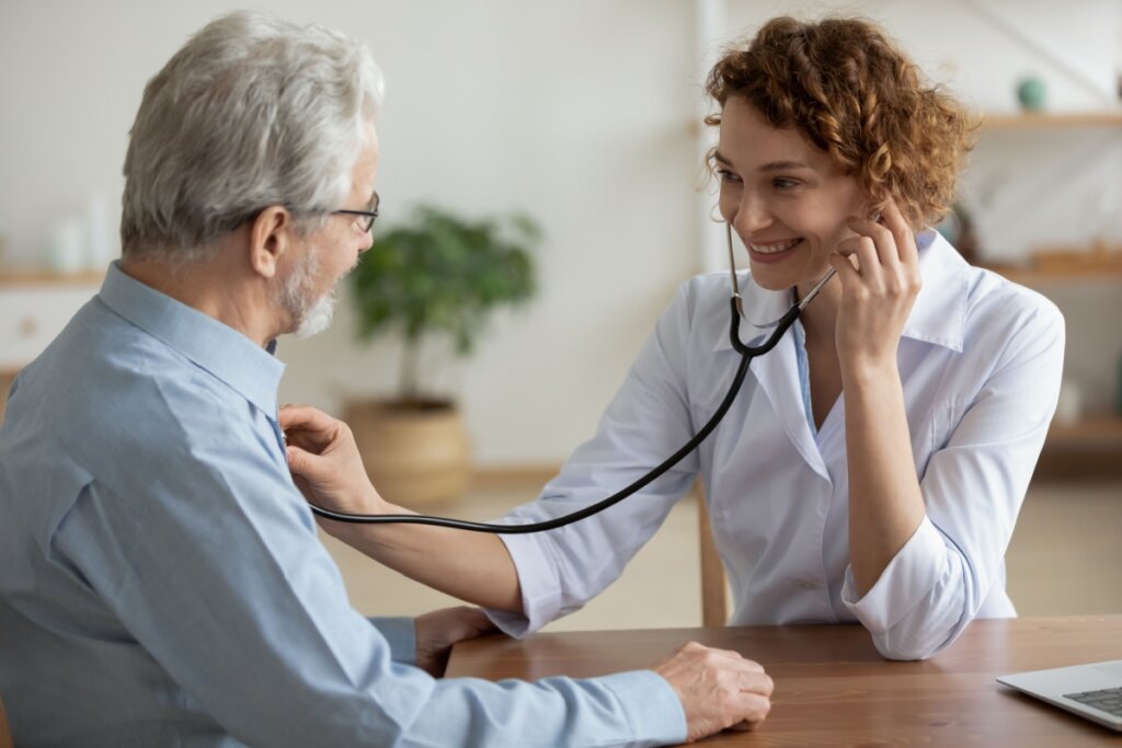 Idoso em consultório sendo avaliado por cardiologista, homem tem o cabelo branco, barba grisalha e está usando óculos e camisa social azul e a médica tem o cabelo curto cacheado e está usando jaleco branco
