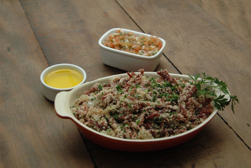 Recipiente branco com farofa com carne assada, cebola e cebolinha em uma mesa de madeira 