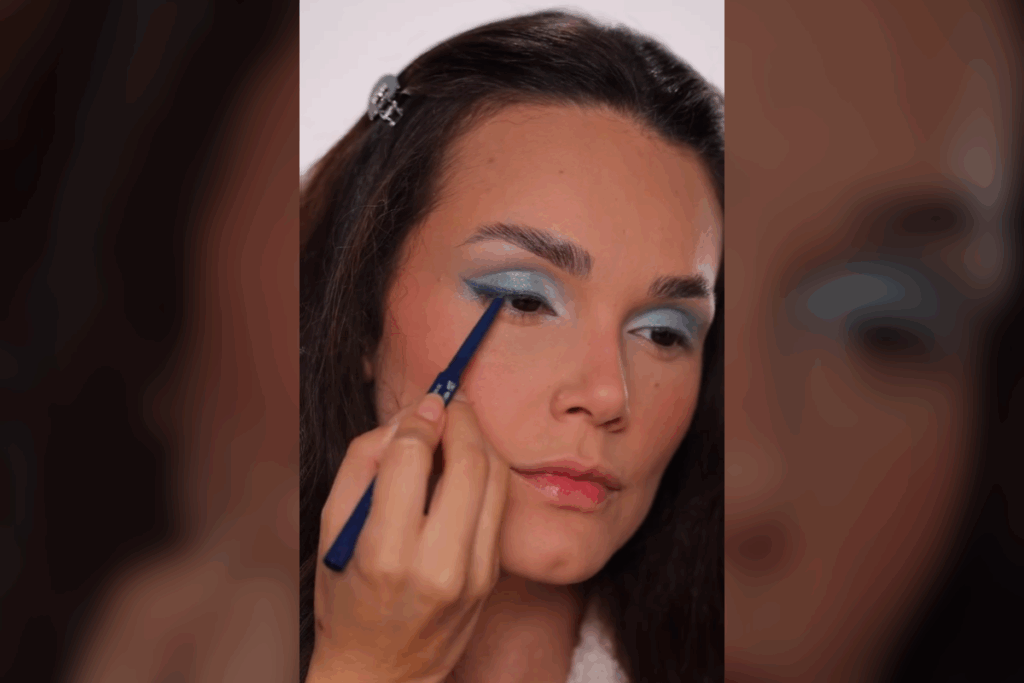 Mulher finalizando a maquiagem dos olhos com um lápis de olho azul-escuro rente aos cílios superiores, criando um leve delineado. A sombra azul-claro já está aplicada em toda a pálpebra, e ela segura o lápis com precisão enquanto olha para baixo.