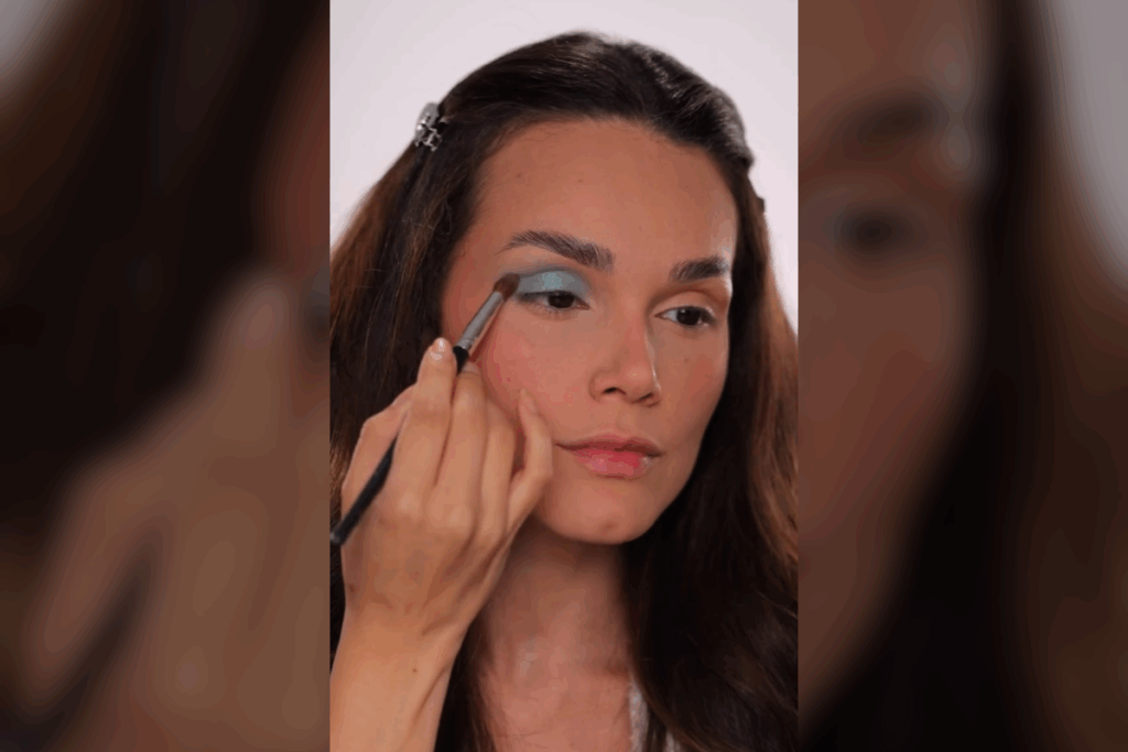 Mulher jovem aplicando sombra azul-claro matte no canto externo da pálpebra superior com um pincel de esfumar pequeno. O acabamento é suave e bem esfumado em direção à sobrancelha. Ela tem cabelos castanhos e o rosto está levemente inclinado.