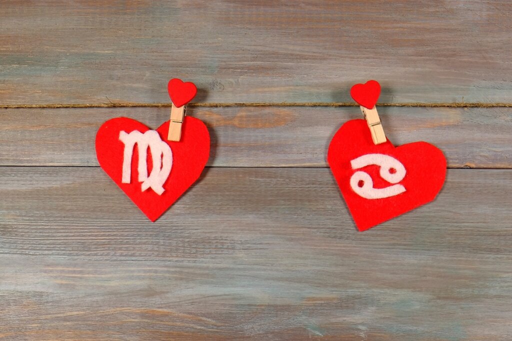 Dois corações de feltro vermelho pendurados em um varal de corda. O coração da esquerda apresenta o símbolo de Virgem e o da direita apresenta o símbolo de Câncer (os dois "69" horizontais), ambos em relevo branco sobre o feltro.