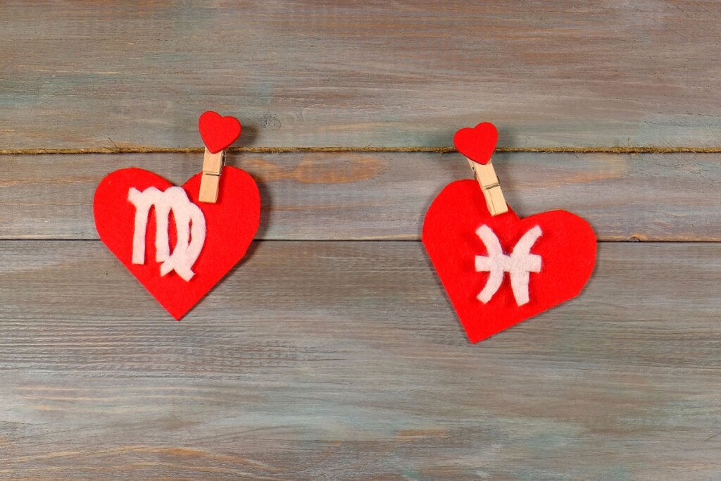 Dois corações de feltro vermelho pendurados por prendedores de madeira em um varal de corda, contra um fundo de madeira rústica. O coração da esquerda exibe o símbolo de Virgem e o da direita exibe o símbolo de Peixes (duas linhas curvas verticais conectadas por uma barra horizontal), ambos em relevo branco.