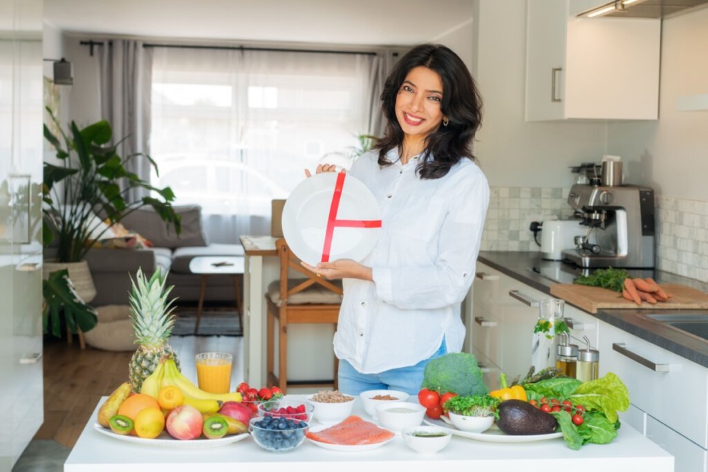 Mulher, em pé em frente a mesa com frutas e vegetais, segando um prato dividido em três partes com fita vermelha