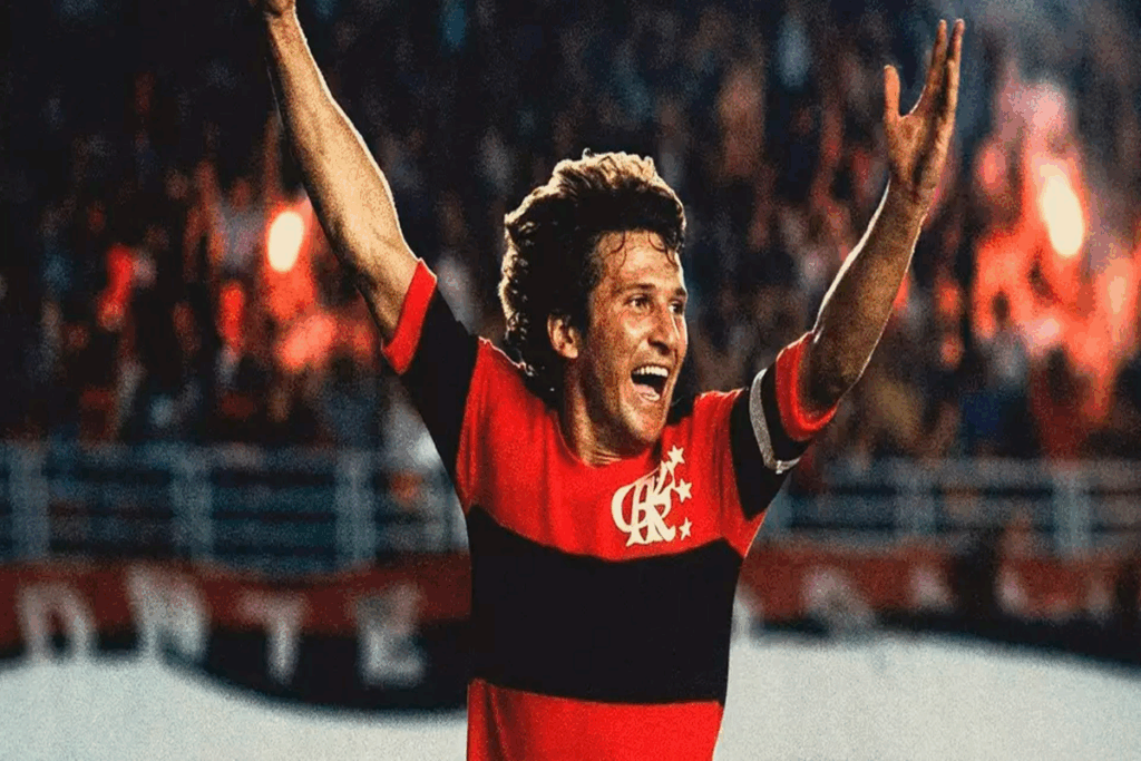 Imagem do jogador de futebol Zico, usando camiseta do Flamengo em jogo com os braços para o alto