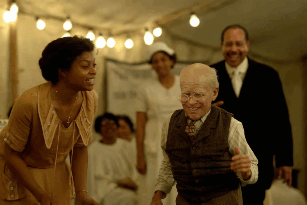 Imagem de uma festa, com uma mulher usando vestido de época bege sorrindo para um menino usando camisa branca e colete marrom e gravata dançando com o rosto envelhecido